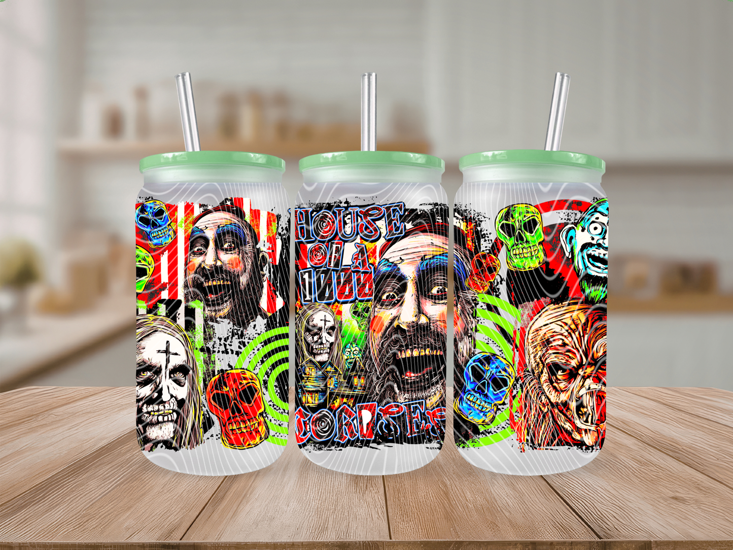 Three clear insulated tumblers with mint green lids and metal straws, decorated with colorful horror-themed artwork including faces, skulls, and words, displayed on a wooden surface with a blurred kitchen background.