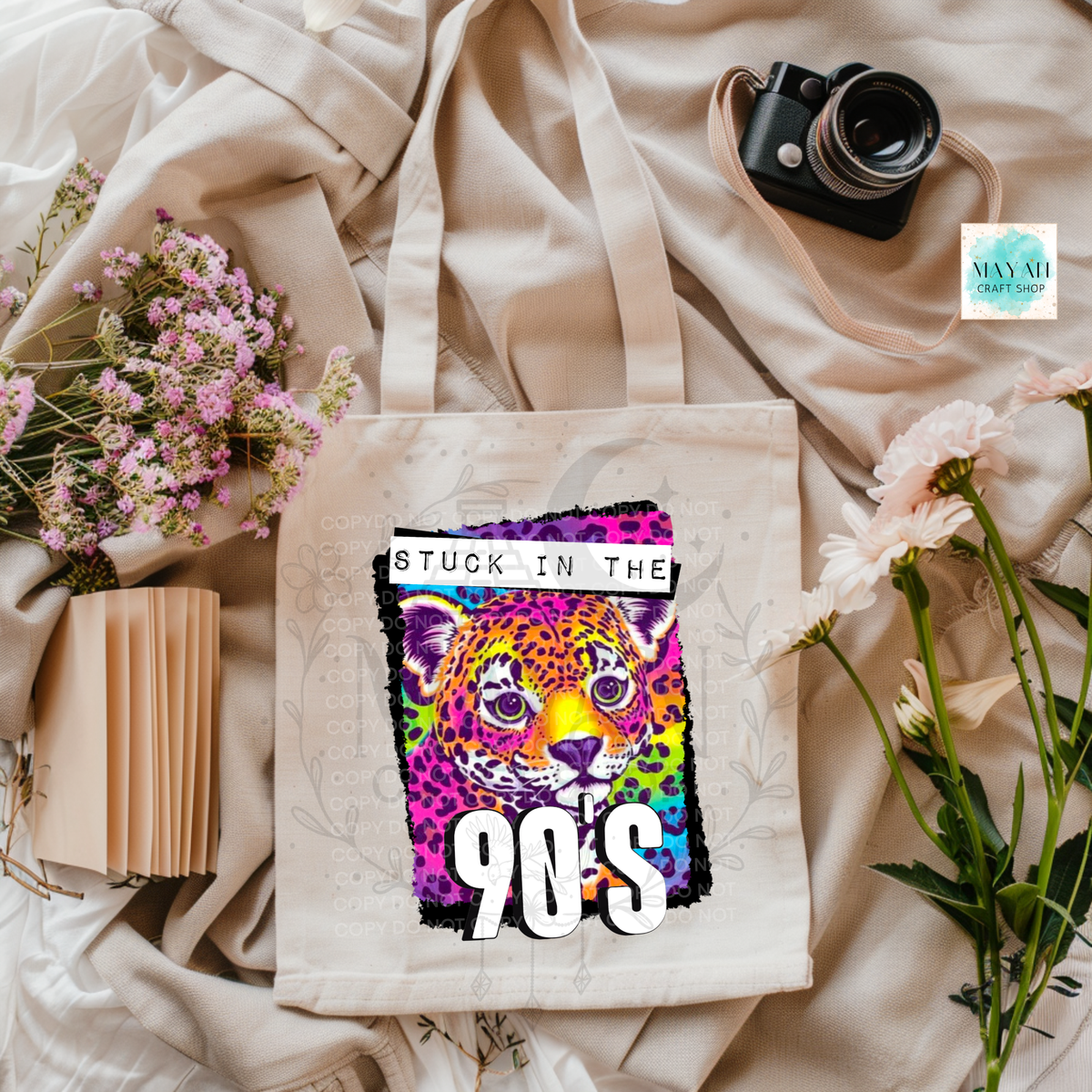 A canvas tote bag with a colorful graphic print of a leopard and the text 'STUCK IN THE 90S' is styled in a flat lay on beige fabric with flowers, a camera, and a notebook.