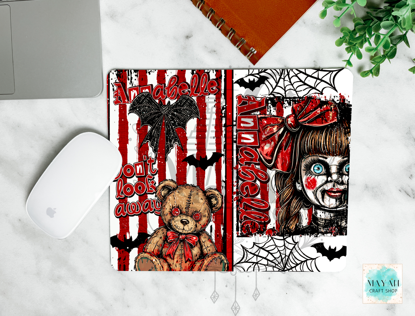 A mouse padt featuring a split Halloween-themed design, shown in a styled flat-lay on a white marble surface next to a laptop, mouse, notebook, and plant. The left half has red and white vertical stripes with a teddy bear, bats, and a text.