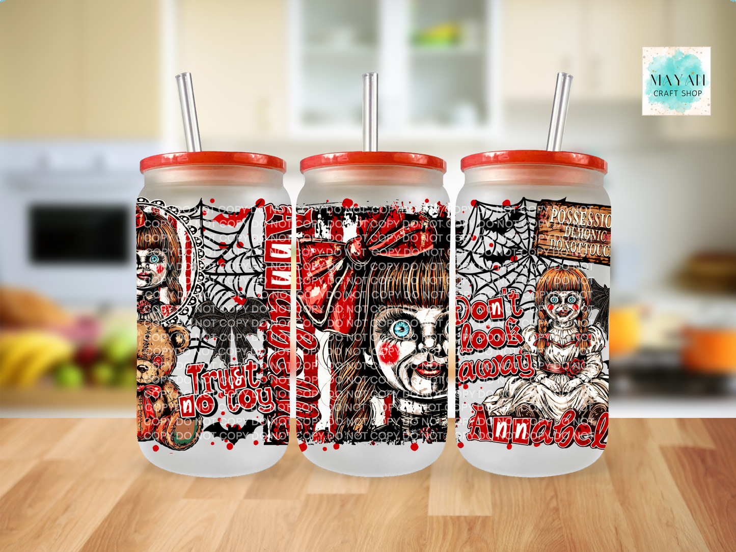 Three frosted glass tumblers with red lids and clear straws, featuring horror-themed illustrated designs in red, black, and white, displayed on a wooden surface with a blurred kitchen background.