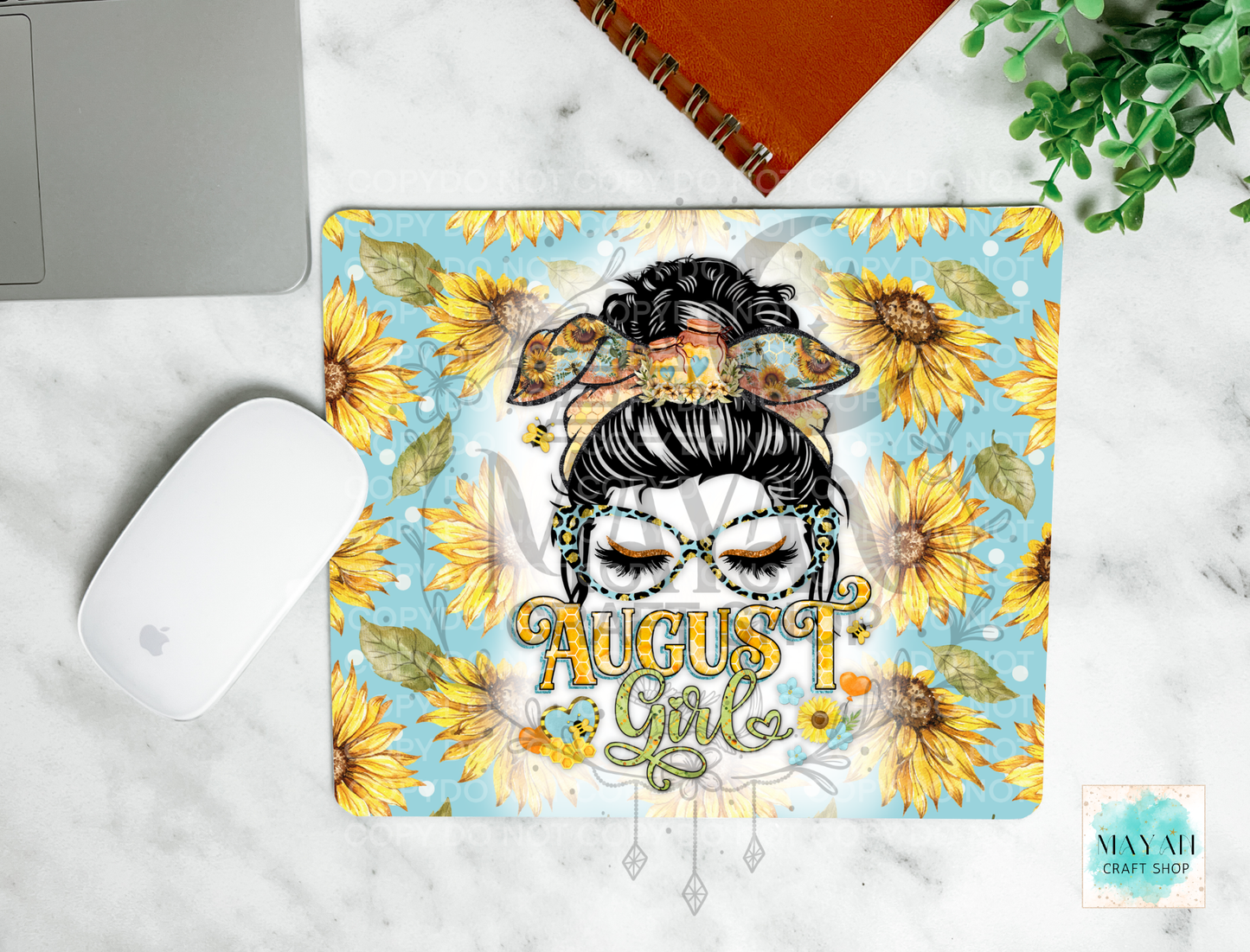 A square mousepad with a colorful 'August Girl' and sunflower design, shown in a flat-lay desk setting on a white marble surface next to a laptop, mouse, notebook, and plant.