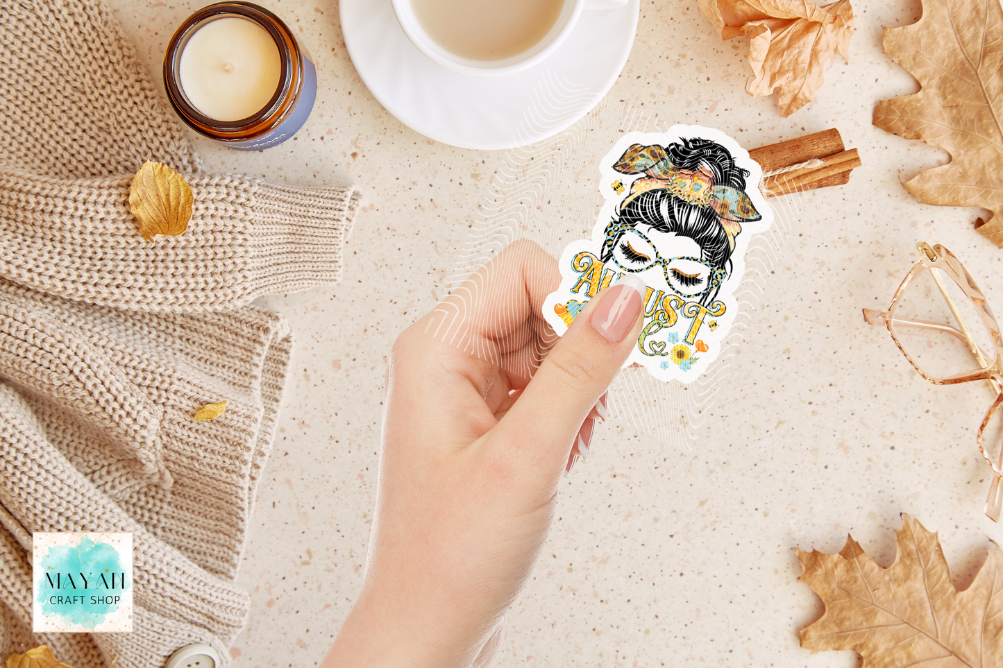 A hand holds a die-cut sticker featuring an illustrated woman with closed eyes, floral details. The scene is styled with a beige knit sweater, a candle, a cup of coffee, cinnamon sticks, glasses, and dried leaves.