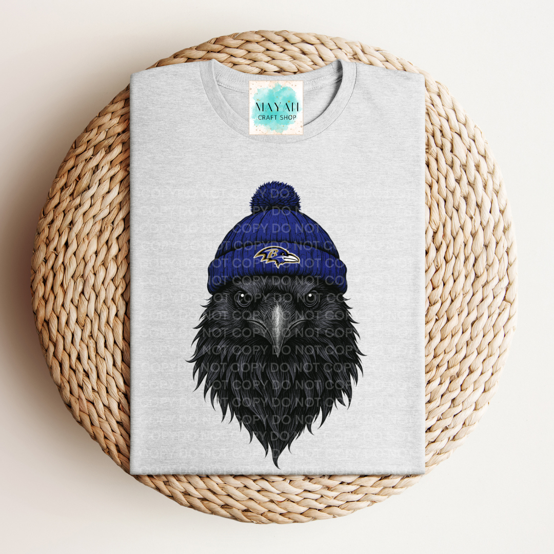 Gray t-shirt with a black crow wearing a blue beanie on a woven mat