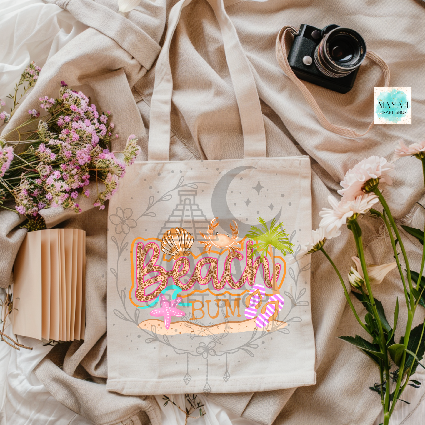 Tote bag with decorative text and graphics on a soft fabric background with flowers and a camera.