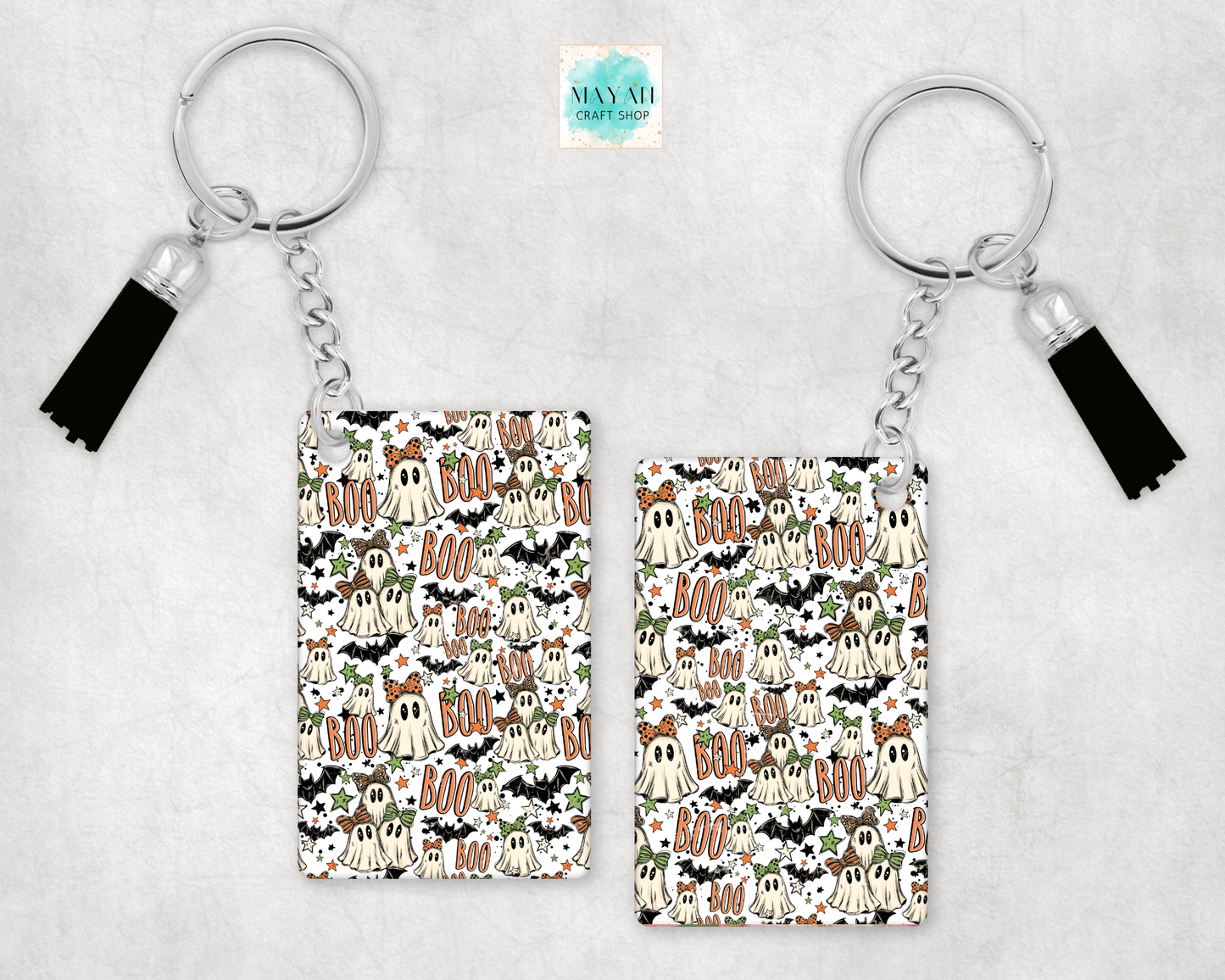Two keychains with black tassels, featuring a repeating Halloween pattern of ghosts, bats, pumpkins, and 'BOO' text on a white background, displayed on a light gray textured surface.
