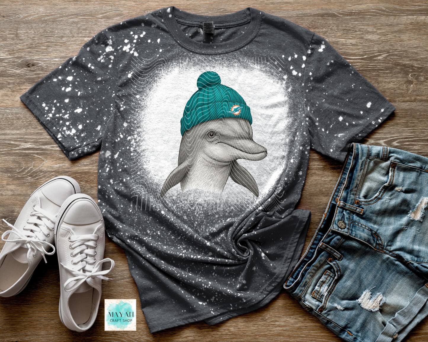 Gray t-shirt with a dolphin wearing a turquoise beanie, paired with white sneakers and blue jeans on a wooden surface.