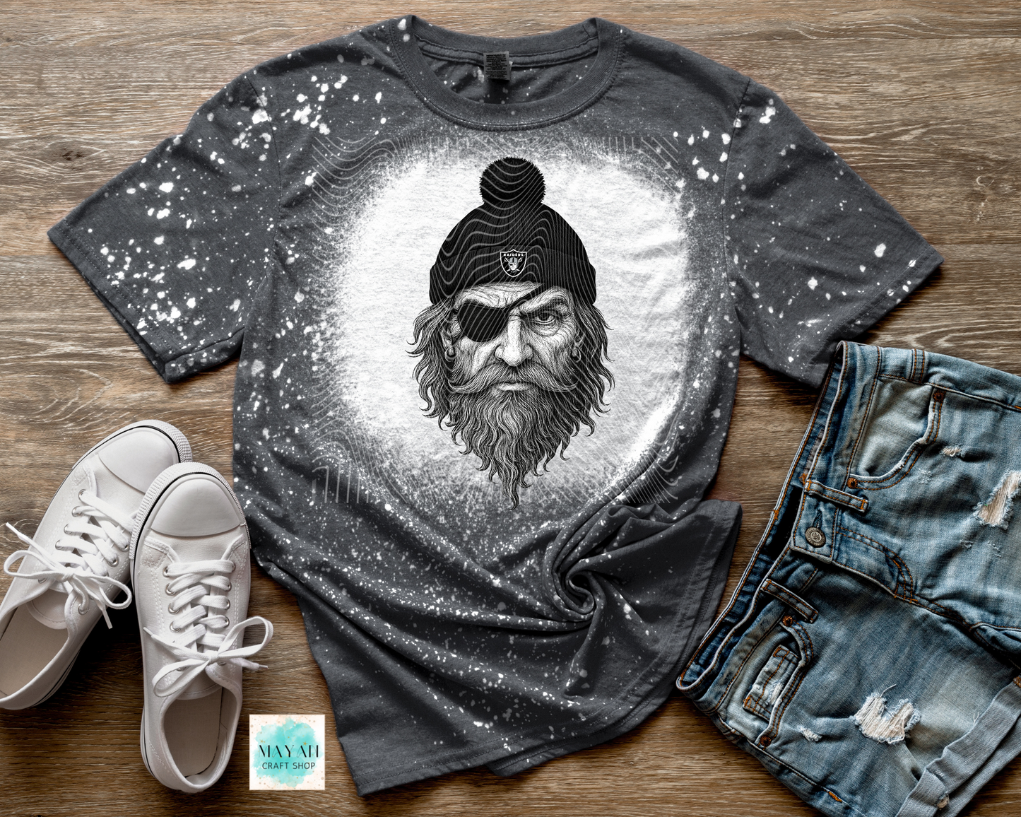 Gray t-shirt with a graphic design of a bearded man wearing a hat and eye patch, paired with white sneakers and blue jeans on a wooden surface.
