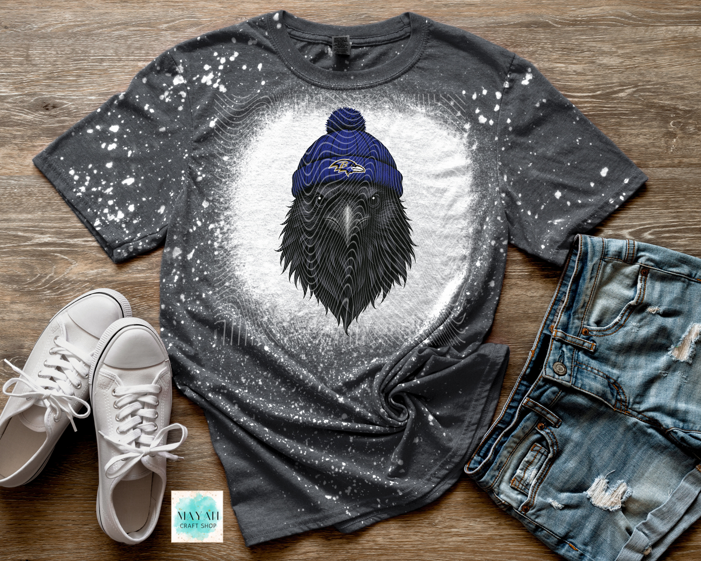 Gray t-shirt with a black crow wearing a blue beanie on a wooden surface with white sneakers and jeans.