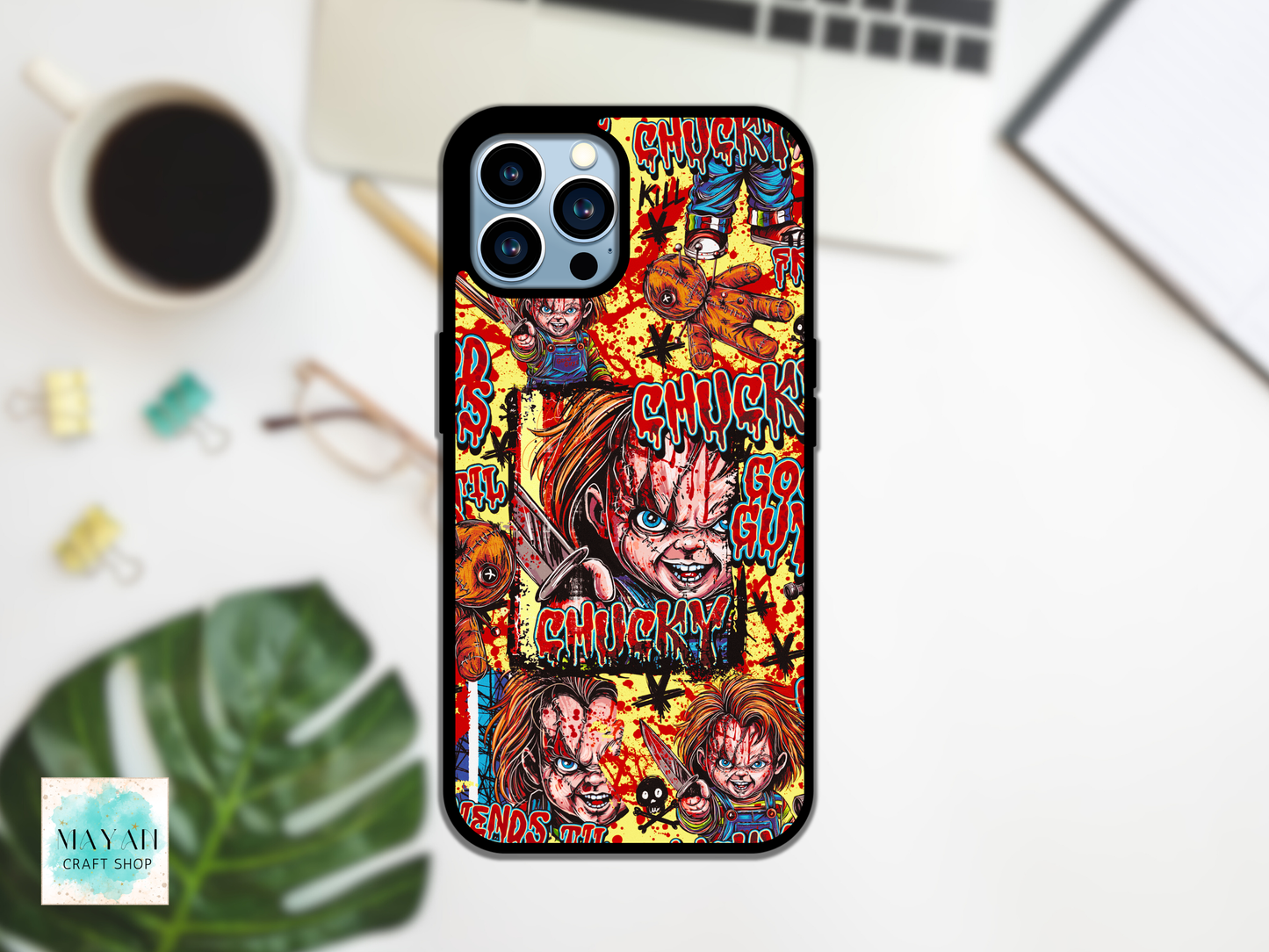 A phone case featuring a colorful collage print, displayed on a white desk with a laptop, coffee cup, and green leaf in the background.