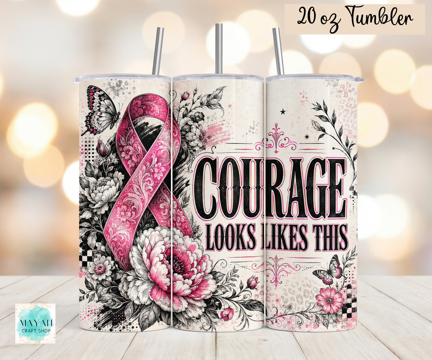 Three 20 oz tumblers with metal straws displayed side-by-side on a light wood surface against a warm bokeh background. The tumblers feature a floral and ribbon design with a pink awareness ribbon, black and white flowers, butterflies, and the text 'COURAGE LOOKS LIKES THIS'.