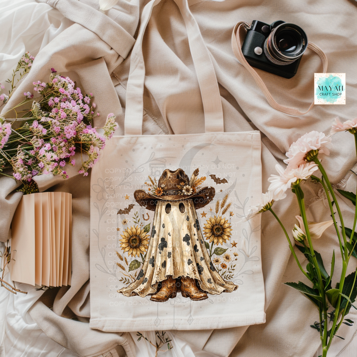 A cream canvas tote bag with a western ghost illustration, styled in a flat lay with flowers, a camera, and a notebook on a beige fabric background.