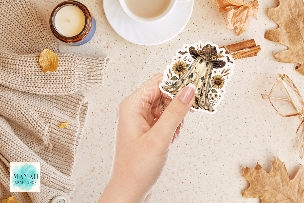 A hand holds a die-cut sticker featuring a vintage-style illustration of a woman with sunflowers and a butterfly, set against a cozy flat-lay background with a cup of coffee, a candle, cinnamon sticks, and autumn leaves.