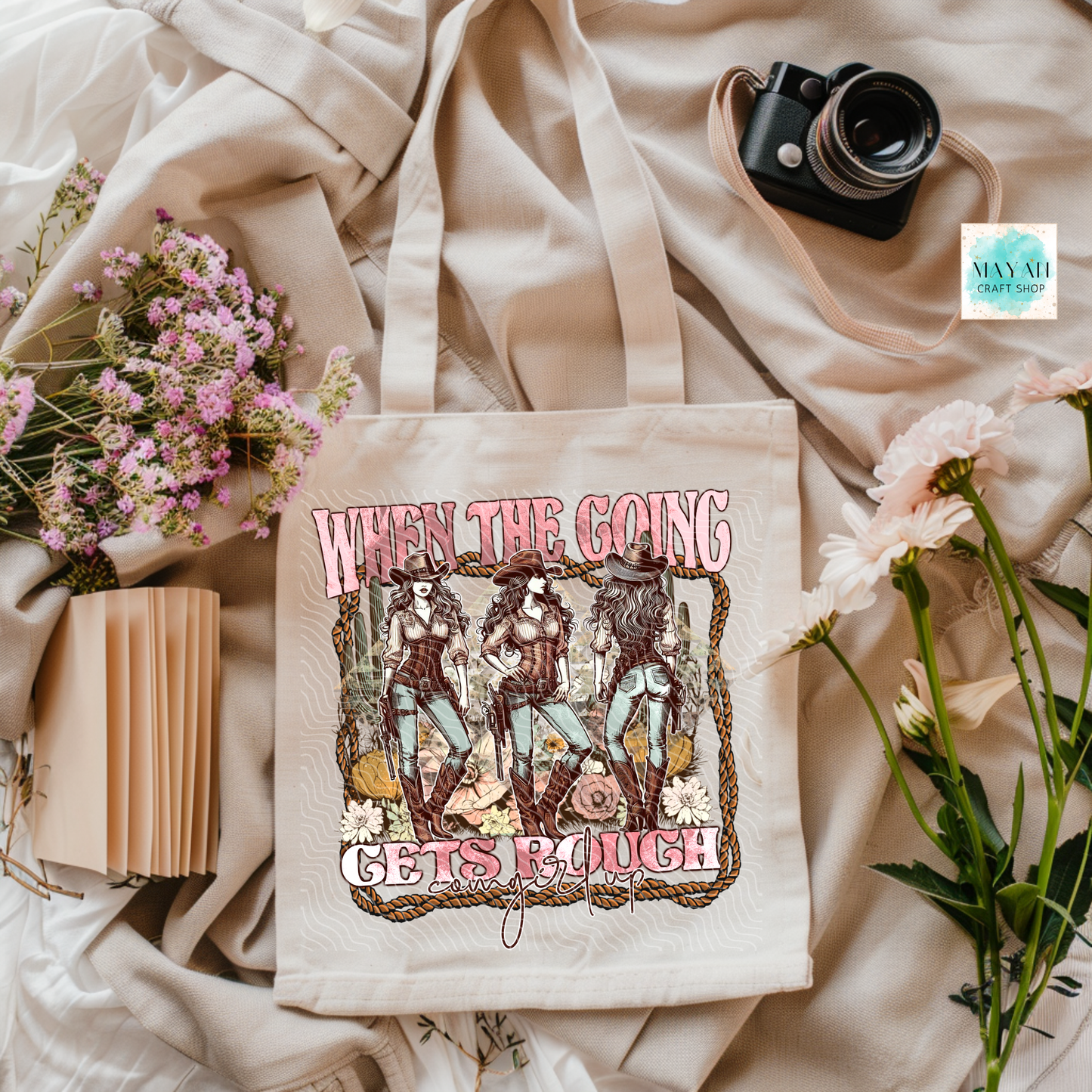 Tote bag with graphic design and text on a soft fabric background with flowers and a camera.