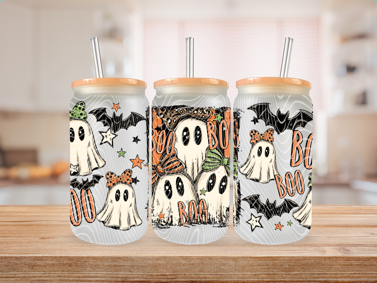 Three clear Halloween-themed tumblers with bamboo lids and metal straws, standing on a wooden table. The tumblers feature cartoon ghost illustrations, bats, stars, and the words 'BOO' in orange, with a softly blurred kitchen background.