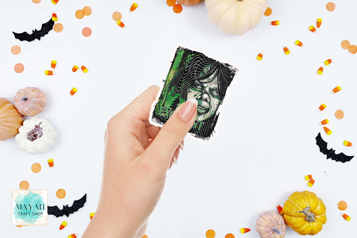 A hand holds a square printed card featuring a spooky green-and-black illustration of a woman's face. The scene is styled with small pumpkins, candy corn, black bat cutouts, and orange confetti on a white background.