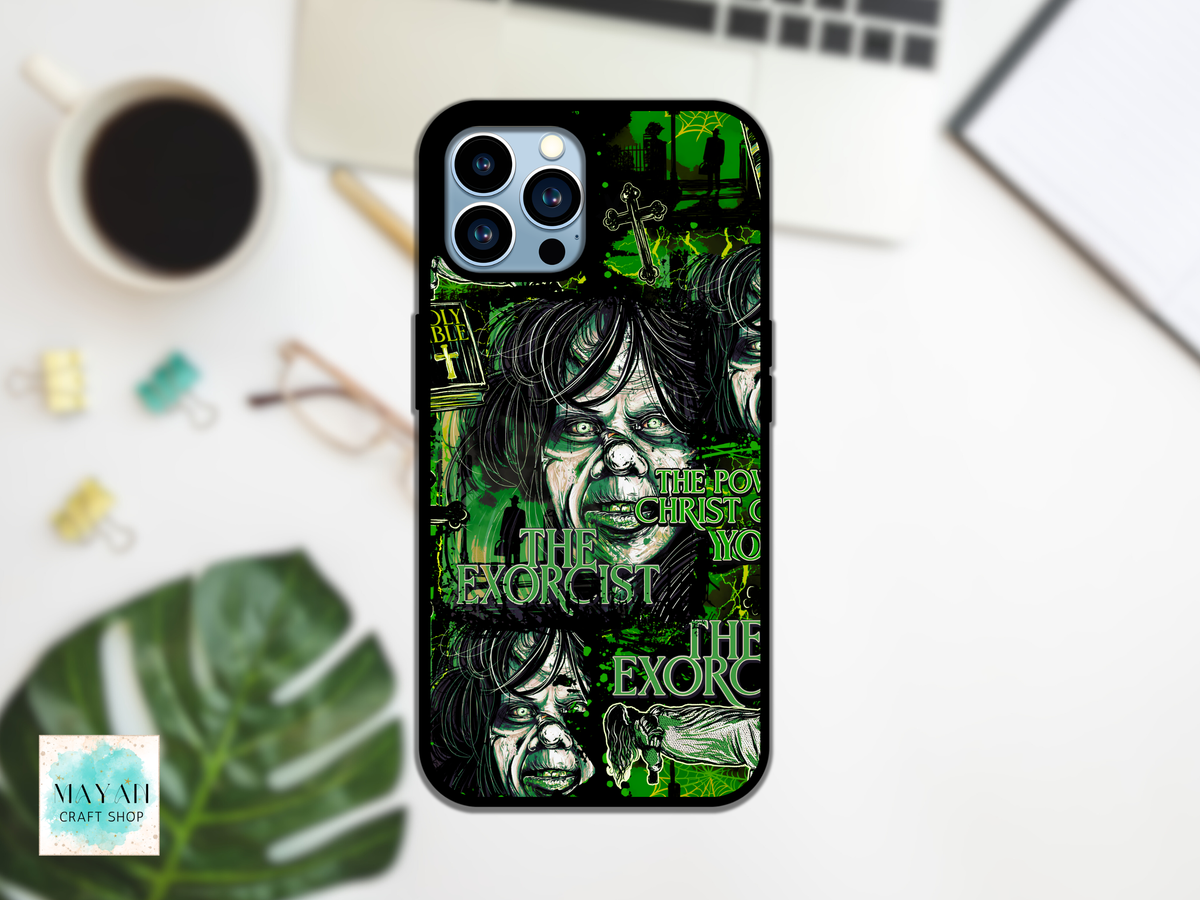 A black phone case with a green and black illustrated design featuring a text and stylized faces, shown on a white desk with a laptop, coffee cup, plant leaf, and binder clips in the background.