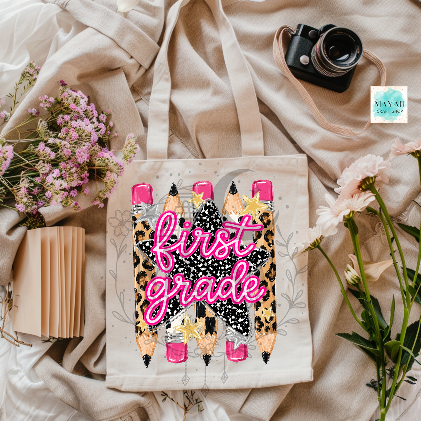 A canvas tote bag with a 'first grade' graphic featuring pink script text over a black-and-white speckled star and pencil illustrations, styled in a flat lay with flowers, a camera, and a notebook on beige fabric.