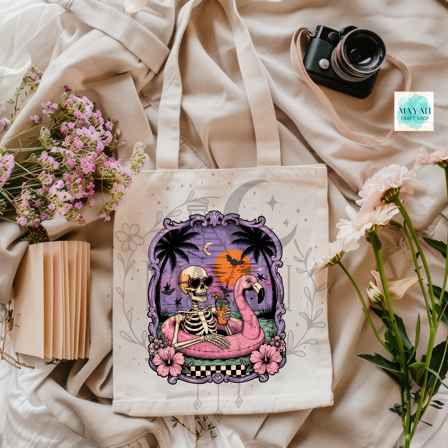 Tote bag with a skeleton and flamingo design on a fabric background with flowers and a camera.