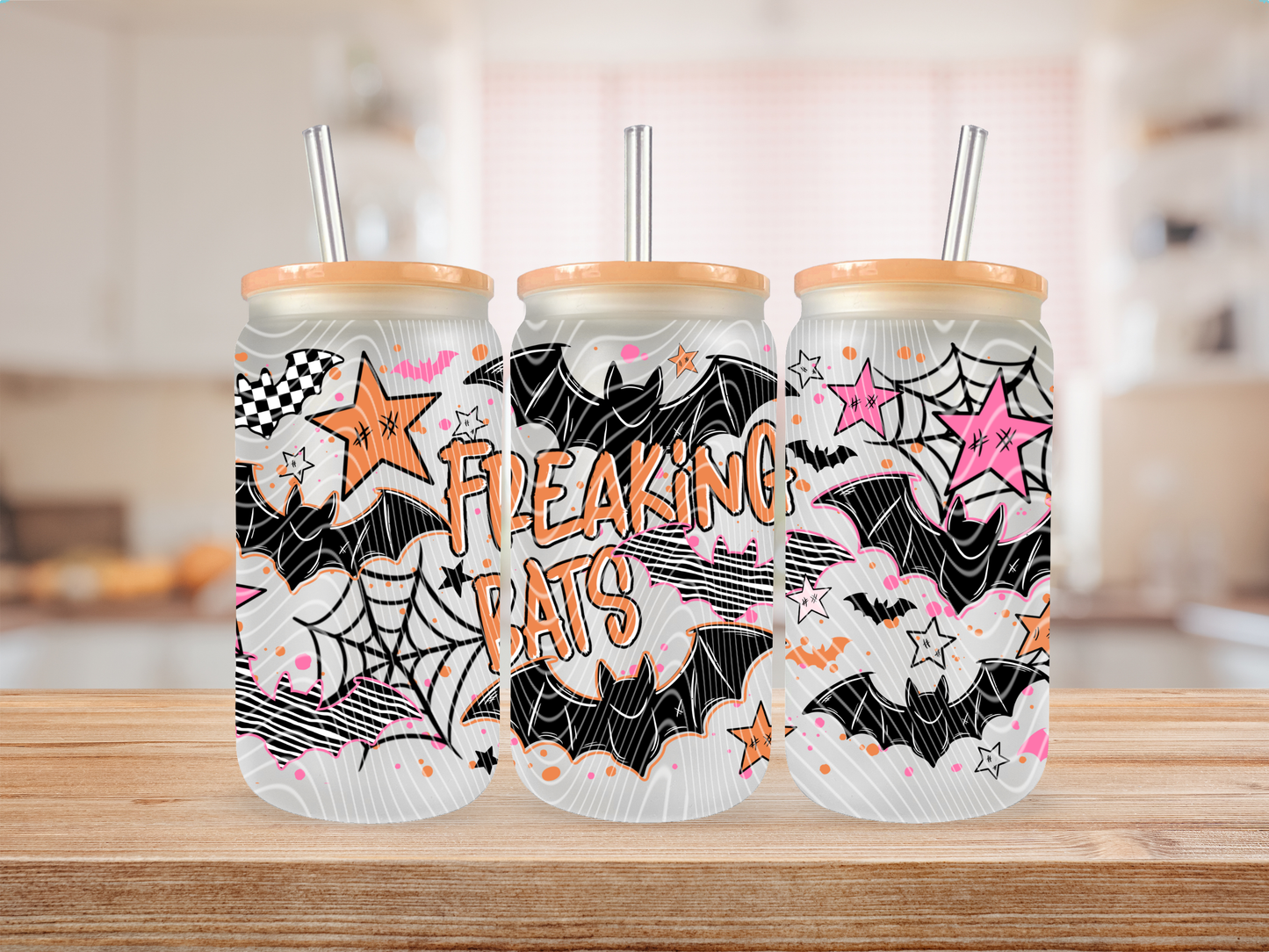 Three frosted insulated tumblers with bamboo lids and metal straws, decorated with a Halloween-themed black, white, orange, and pink pattern featuring bats, spiderwebs, stars, and the words 'FREAKING BATS,' displayed on a wooden surface with a blurred kitchen background.