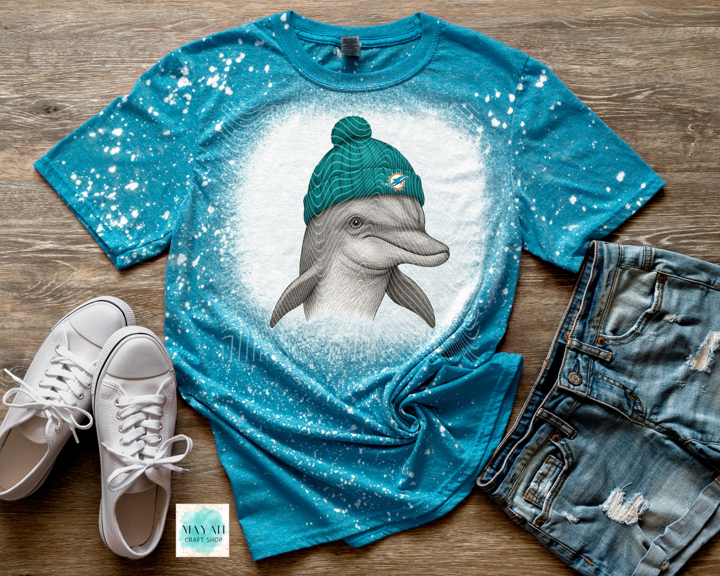 Blue t-shirt with a dolphin wearing a green beanie on a wooden surface with white sneakers and denim shorts.