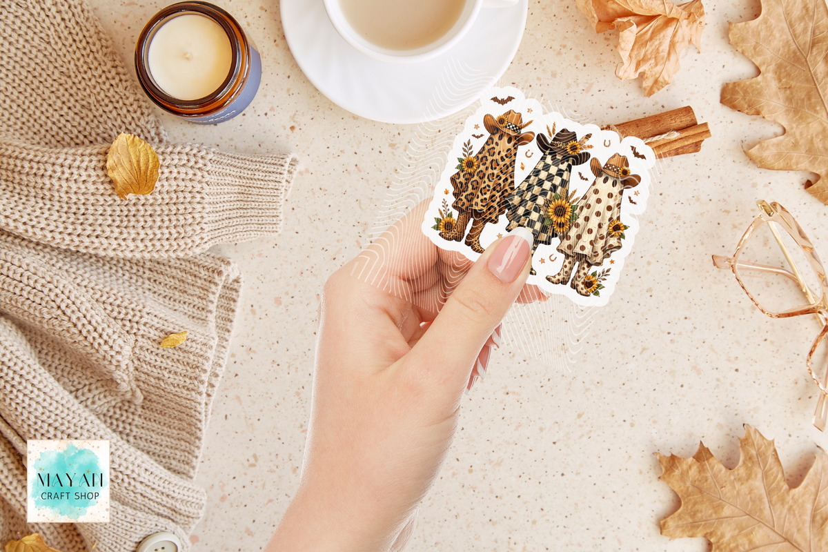 A hand holds a die-cut sticker featuring 3 western ghosts in patterned outfits, surrounded by cozy fall props including a cup of tea, a candle, cinnamon sticks, leaves, and a knitted sweater on a light beige surface.