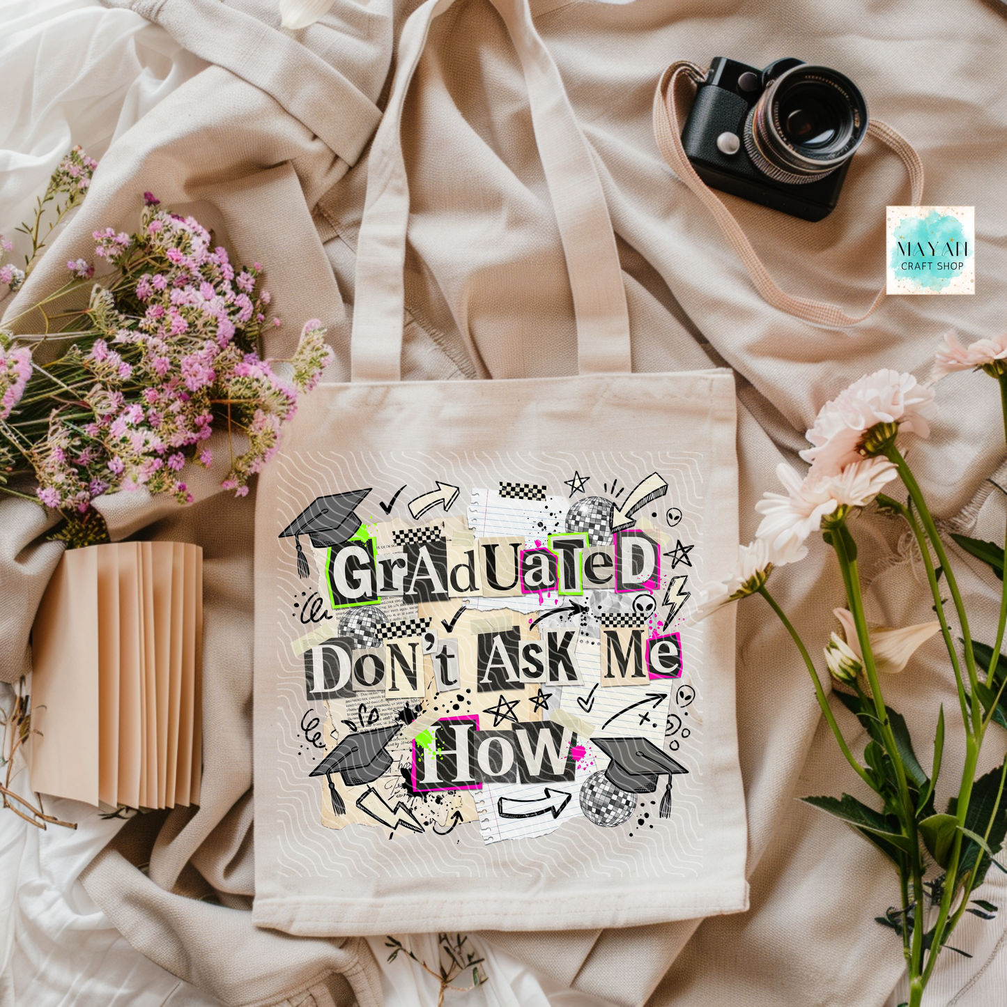 Tote bag with 'Graduated Don't Ask Me How' text on a soft fabric background with flowers and a camera.