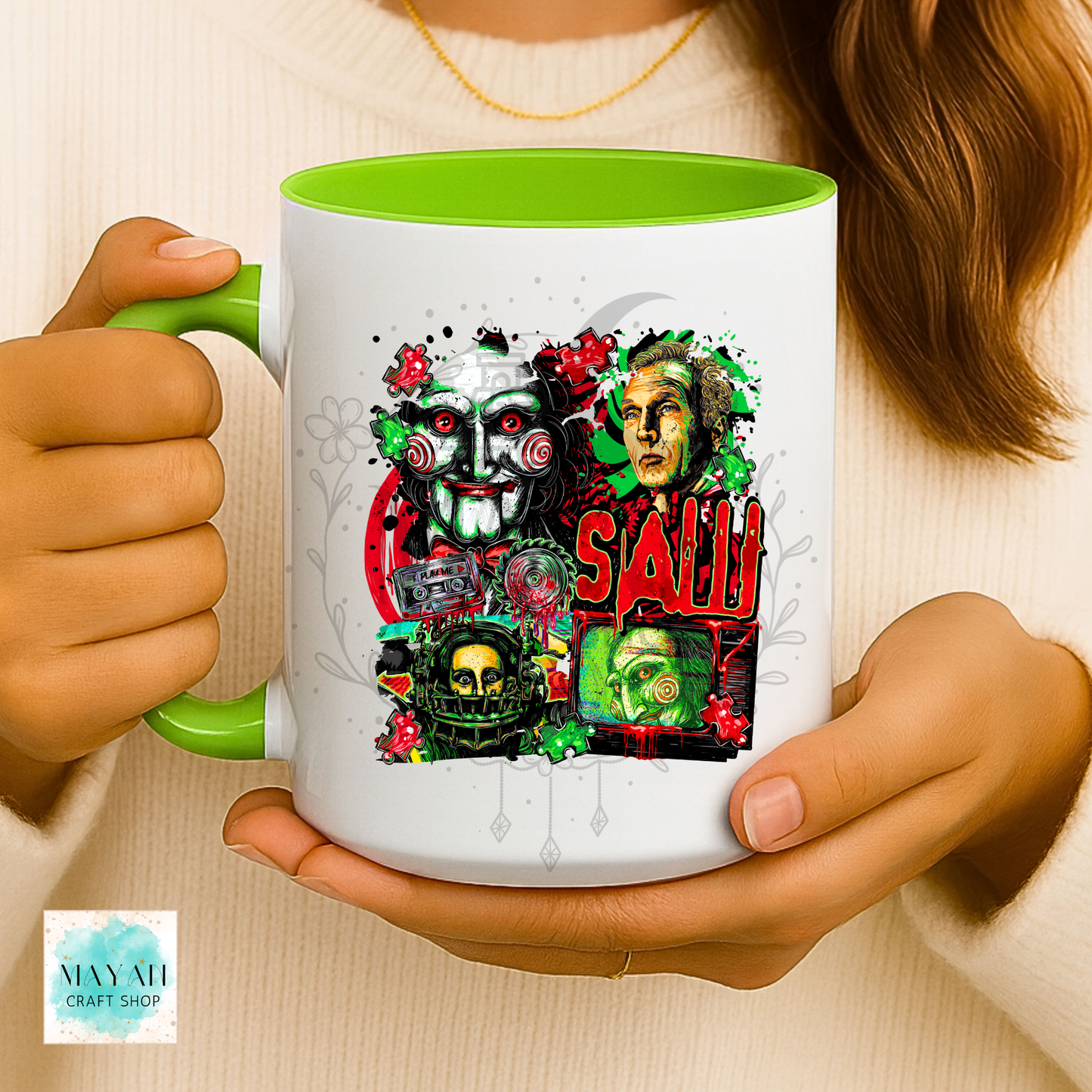A person holding a white ceramic mug with a bright green handle and interior. The mug features a colorful graphic collage of horror movie masks and the a word in red.