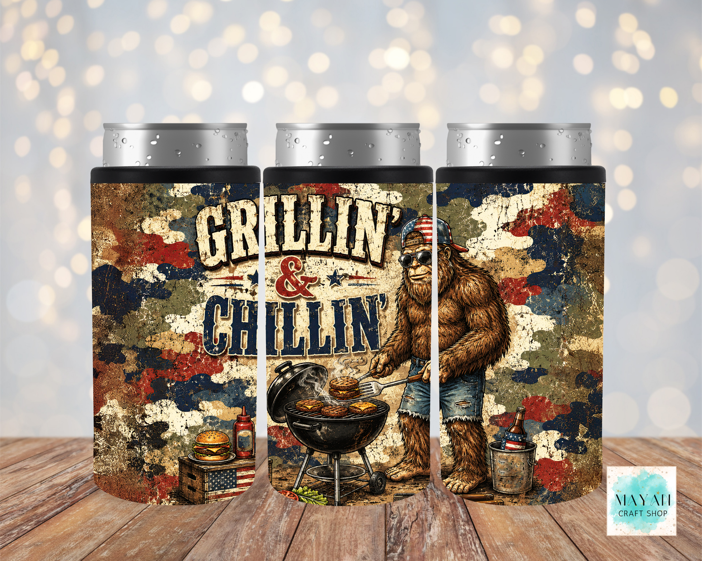 Three cylindrical can cooler with a creature grilling design and 'Grillin' & Chillin'' text on a wooden surface.