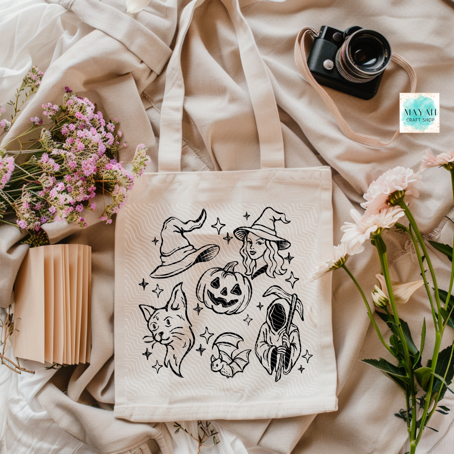 Beige tote bag with Halloween-themed designs on a soft beige fabric background with flowers and a camera.