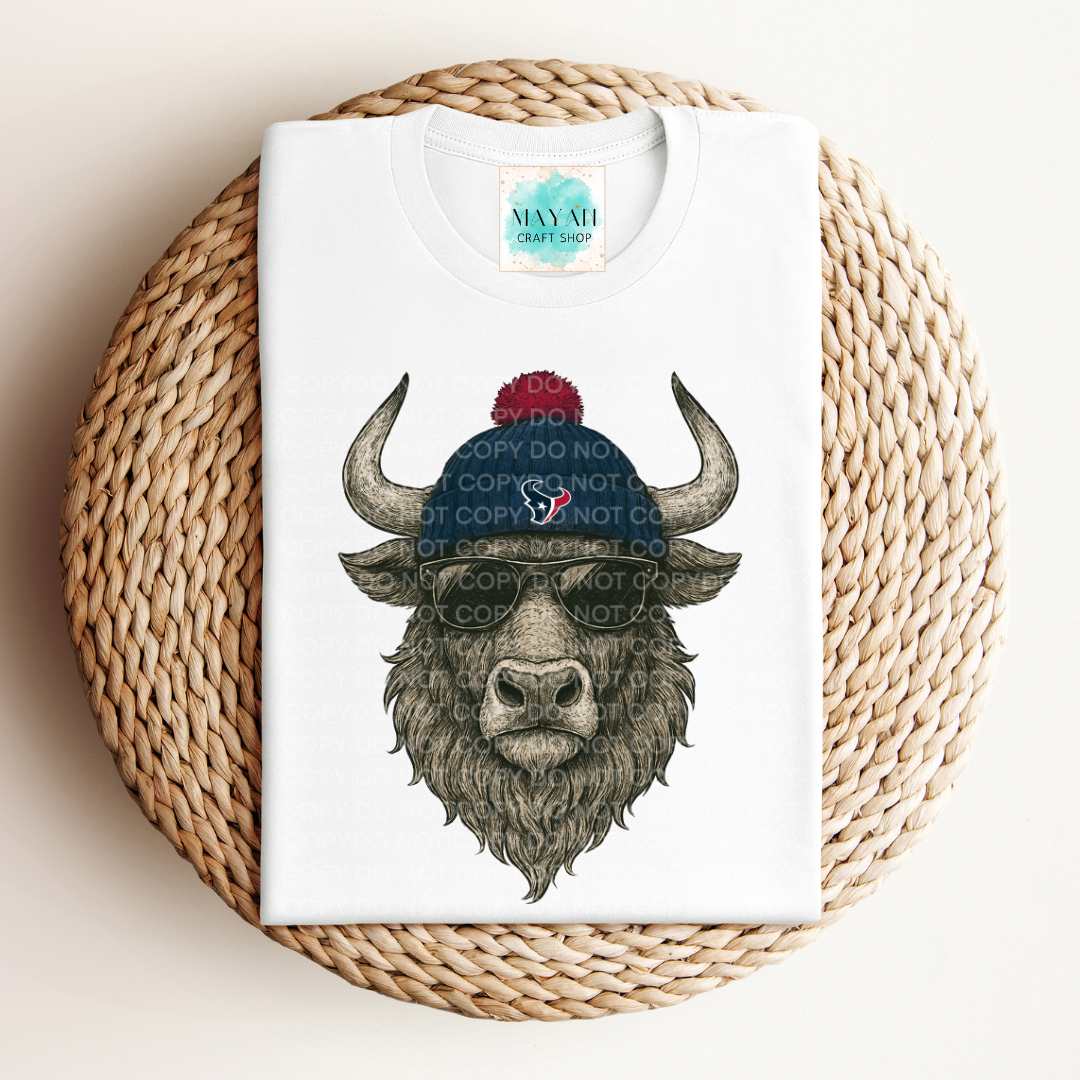 White t-shirt with a graphic of a bison wearing a blue beanie with a red logo, placed on a woven mat.