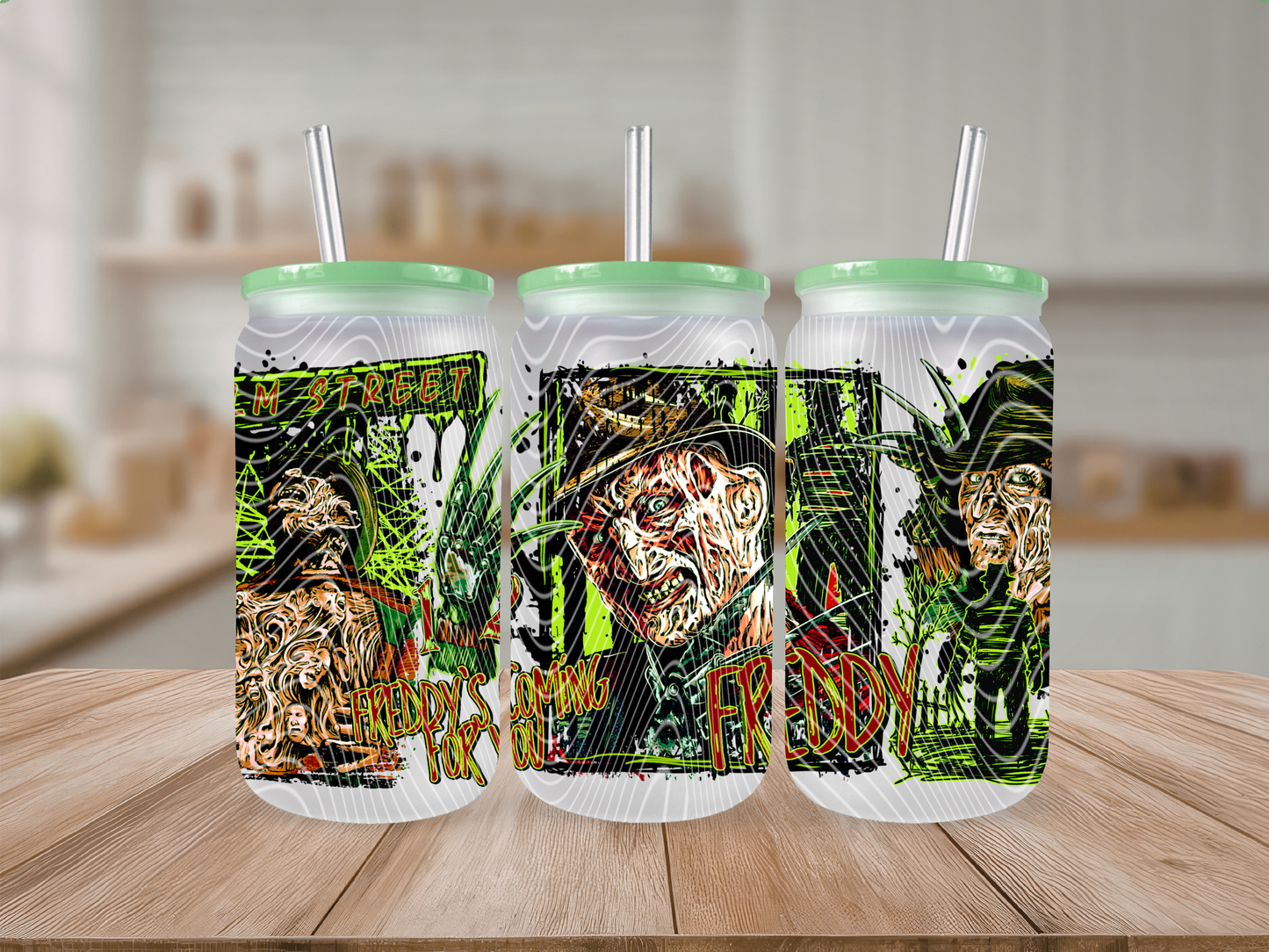 Three frosted insulated tumblers with mint green lids and straws, featuring a horror-themed graphic print, displayed on a wooden table with a blurred kitchen background.