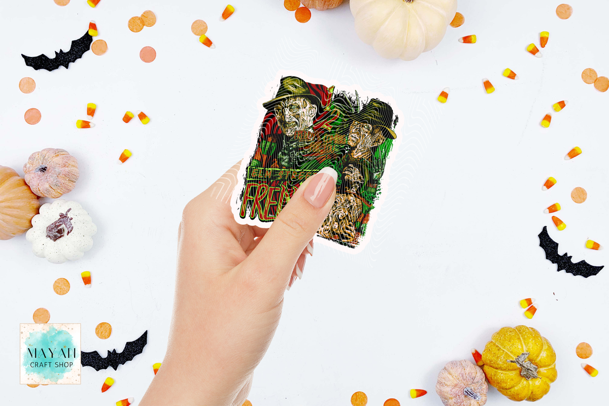 A hand holds a sticker featuring a spooky illustration of three skeletons wearing green hats and scarves, with a text. The background is a white flat-lay styled with mini pumpkins, candy corn, black bat cutouts, and orange confetti.