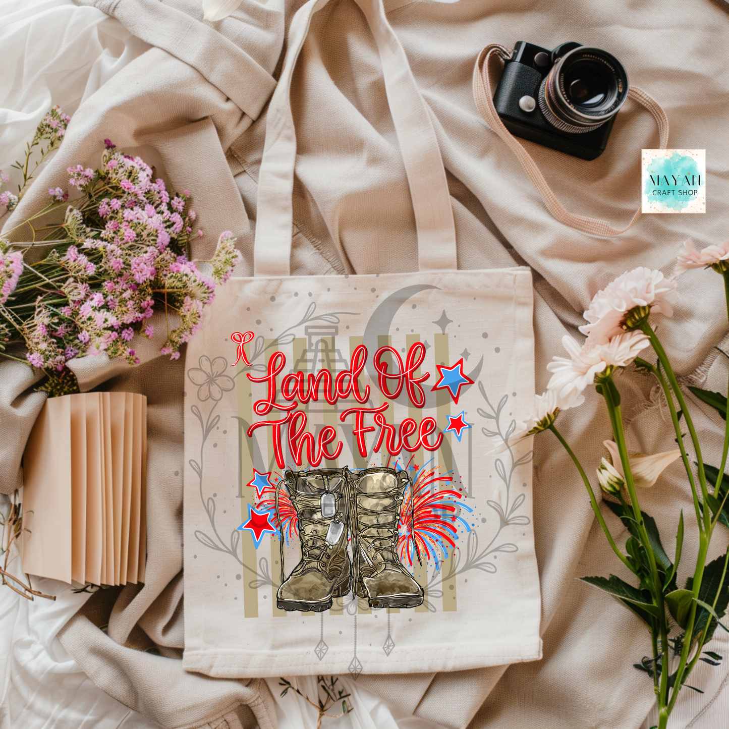 Tote bag with 'Land of the Free' text and boots design on a fabric background with flowers and a camera.
