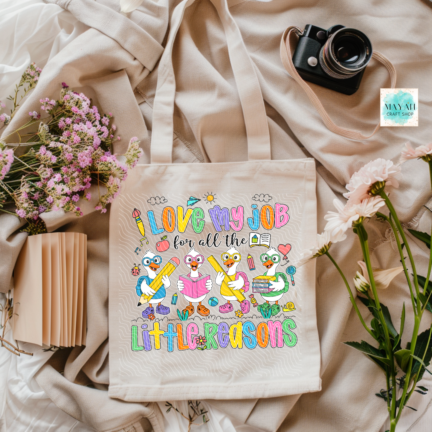 Tote bag with colorful design and text on a soft fabric background with flowers and a camera.