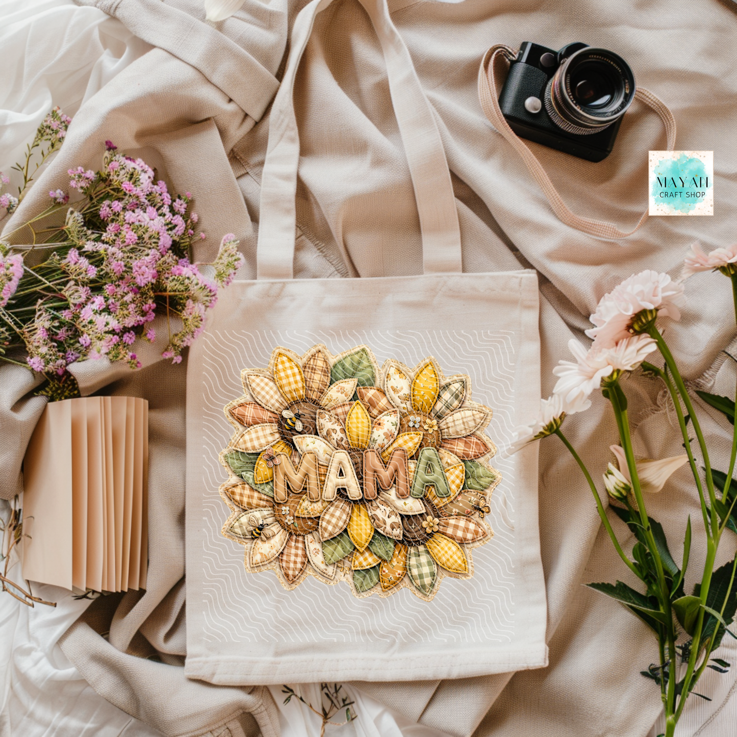 Beige tote bag with 'MAM' design surrounded by flowers and a camera on a light fabric background