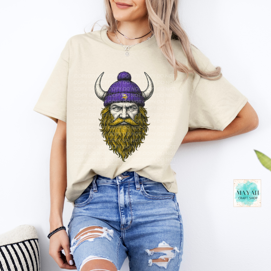 Person wearing a beige t-shirt with a graphic design of a bearded man in a purple beanie, standing against a white background.