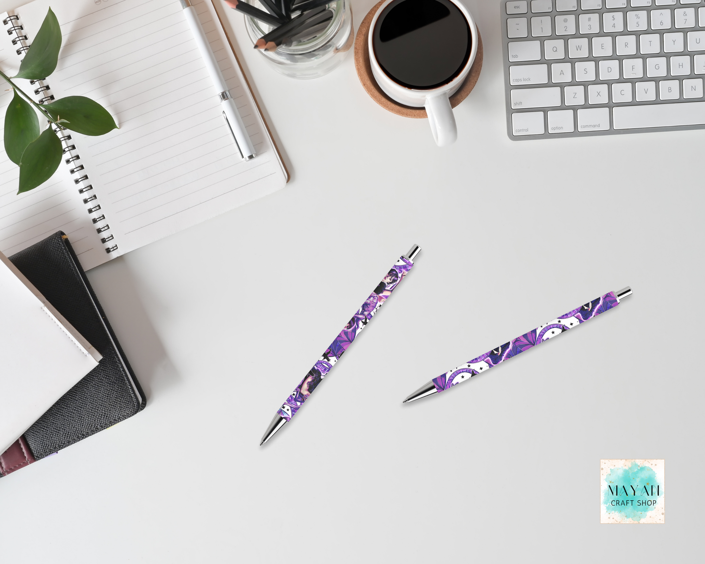 Two purple pens with floral design on a desk with a notebook, coffee cup, and keyboard.