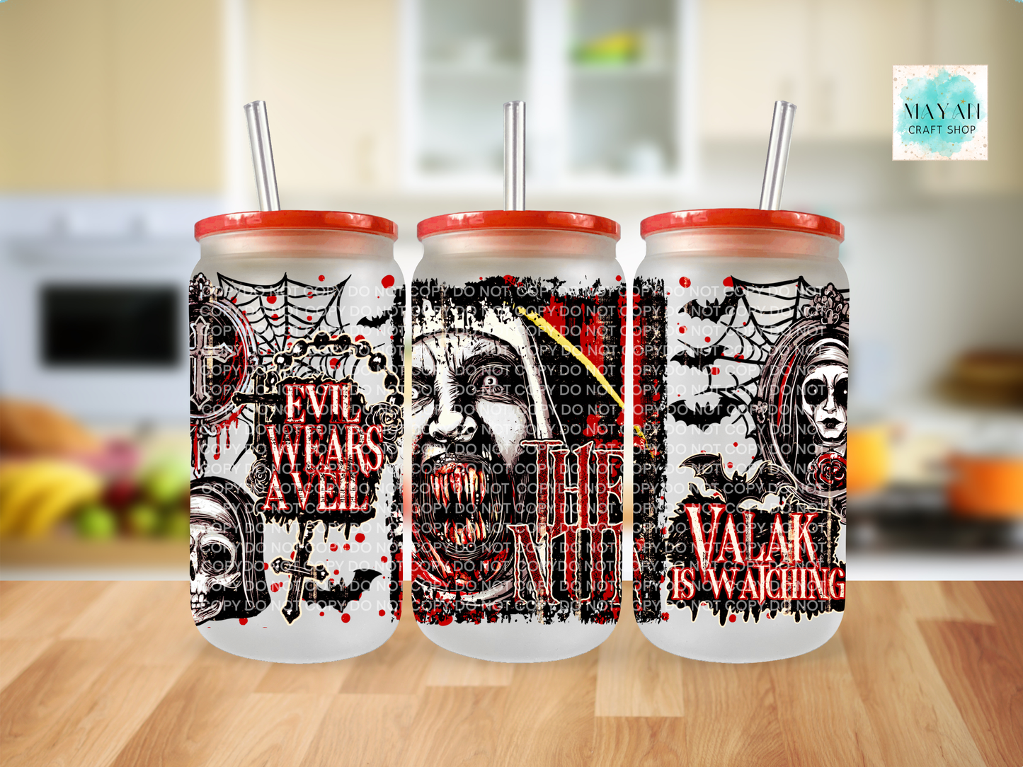 Three Halloween-themed glass tumblers with red lids and clear straws, displayed on a wooden table with a blurred kitchen background. The tumblers feature spooky black and red graphics.
