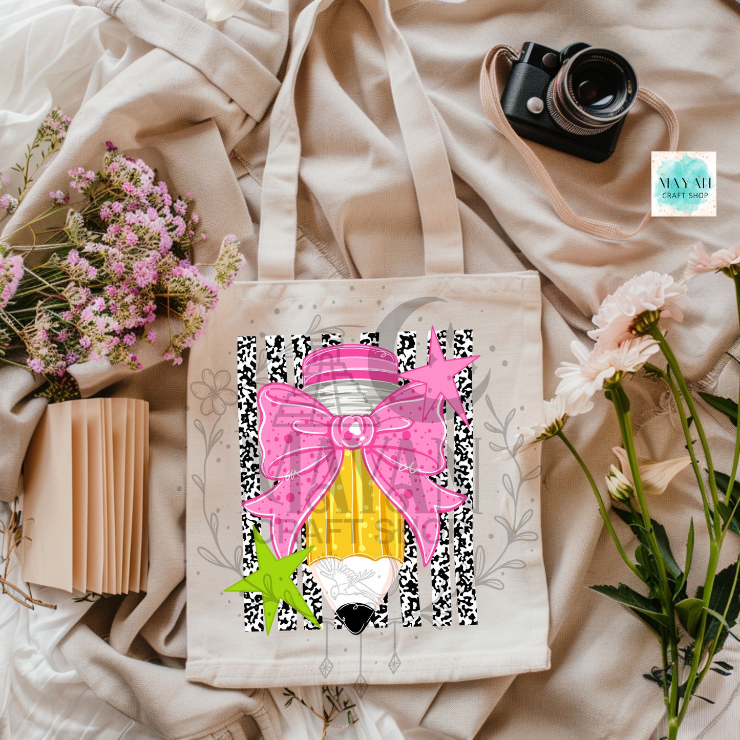 A canvas tote bag with a colorful graphic print of a gnome wearing a large pink bow and pencil, styled in a flat lay with beige fabric, flowers, a book, and a camera.