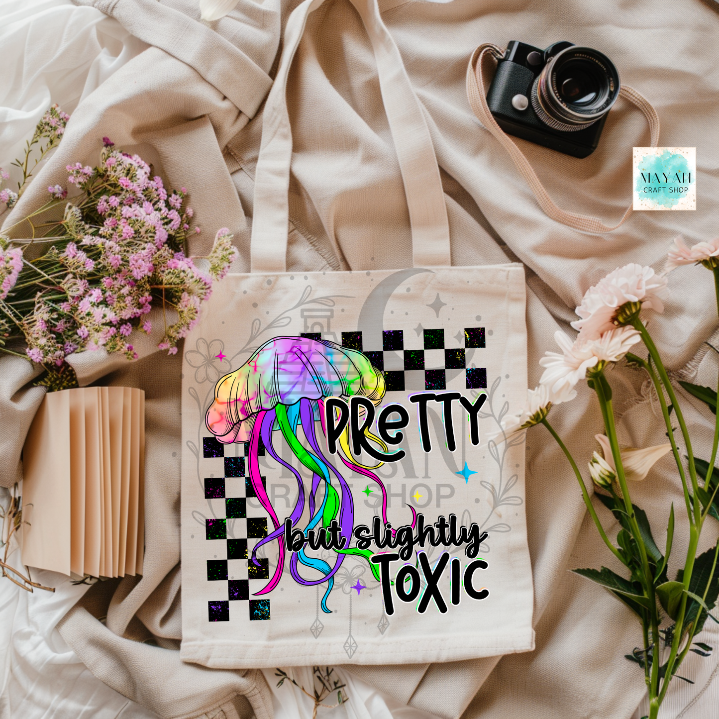 Tote bag with colorful jellyfish design and text on a soft fabric background with flowers and a camera.