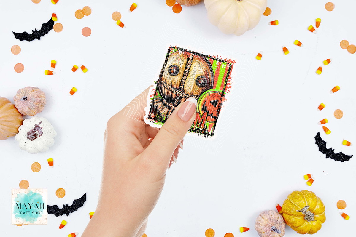 A hand holds a small square Halloween-themed sticker or patch featuring a stitched jack-o'-lantern and a green witch hat, surrounded by pumpkins, candy corn, and black bat cutouts on a white background.