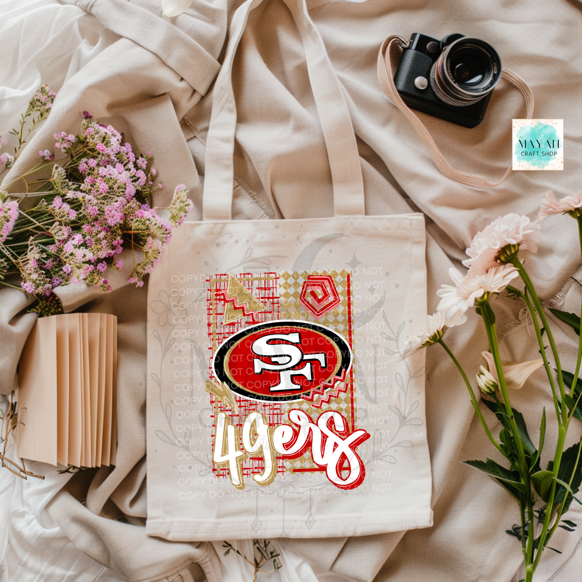 A cream canvas tote bag featuring a football team logo graphic print, styled in a flat-lay arrangement with a vintage camera, pink flowers, a small notebook, and draped beige fabric.