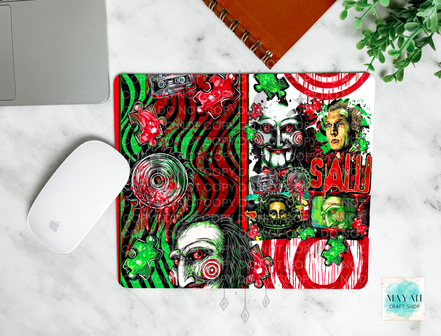 A mouse pad featuring a highly detailed, colorful horror-themed collage design inspired by a horror movie, styled in a flat-lay on a white marble surface next to a white computer mouse and an orange spiral notebook.