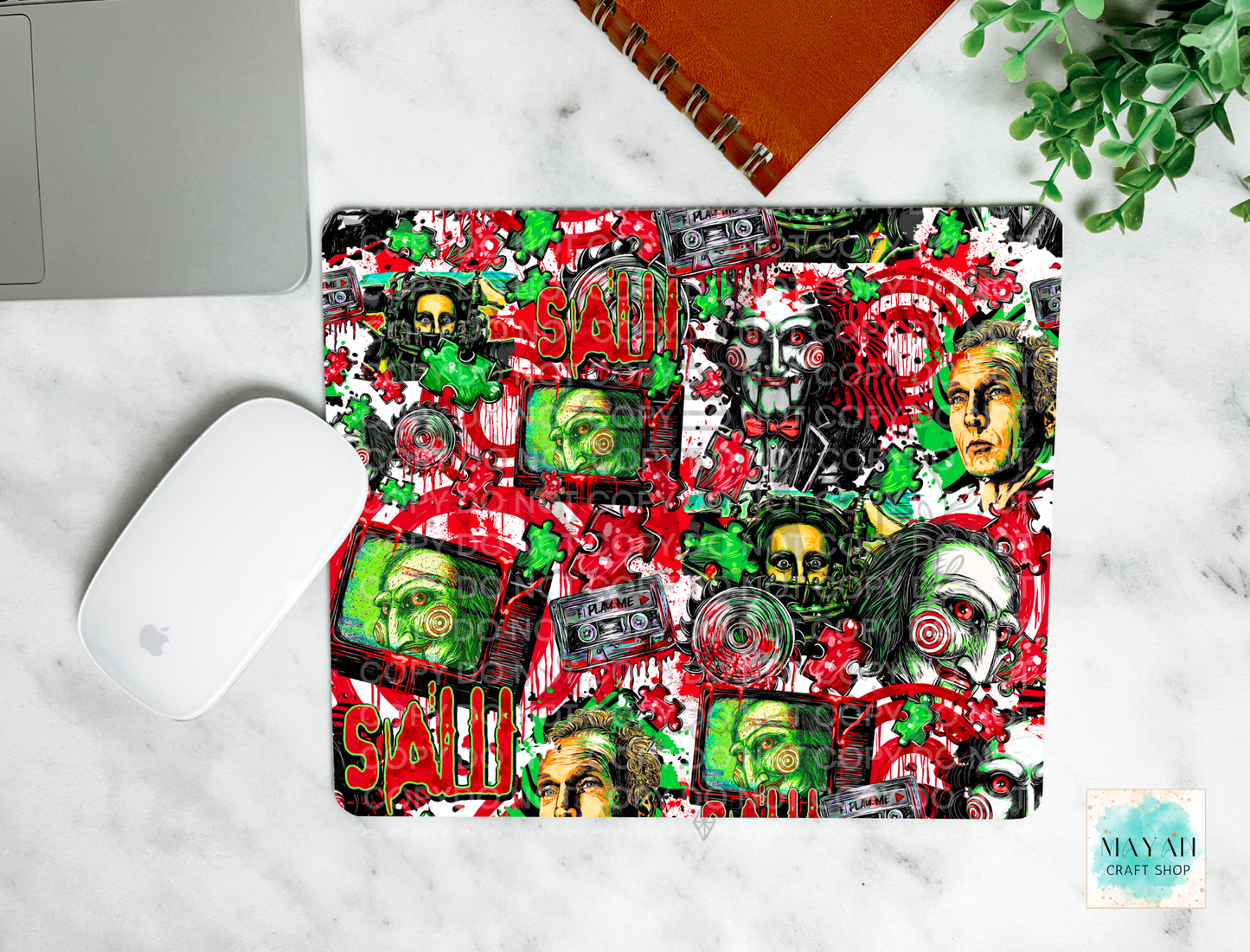 A mouse pad with a vibrant, multicolored collage print featuring the red blood, cassette tapes, skulls, and faces, shown in a flat-lay setup on a white marble desk alongside a laptop, mouse, notebook, and plant.