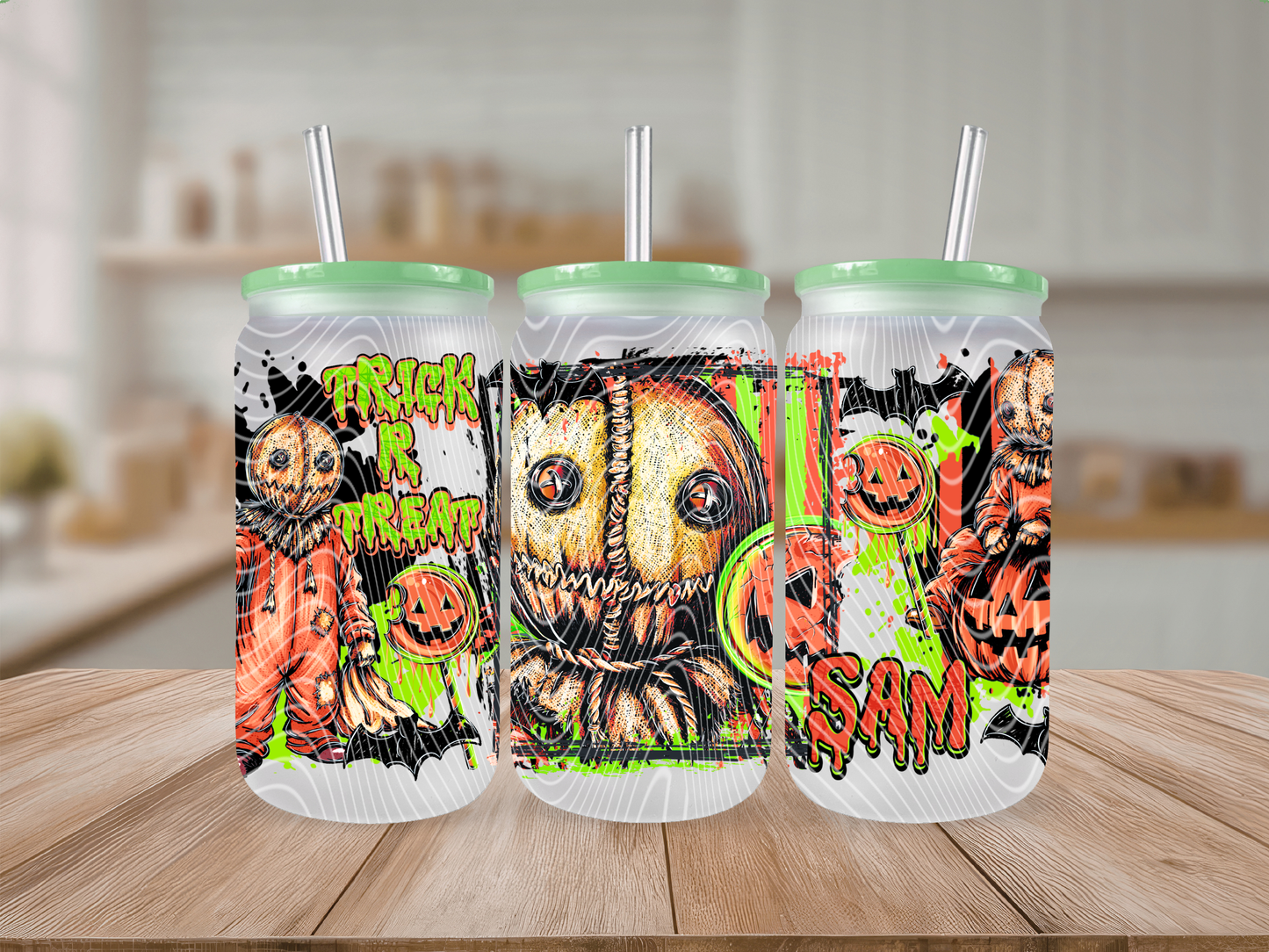 Three frosted glass cans with mint green lids and straws, printed with a Halloween-themed horror design featuring a stitched scarecrow face, pumpkins, displayed on a wooden surface with a blurred kitchen background.