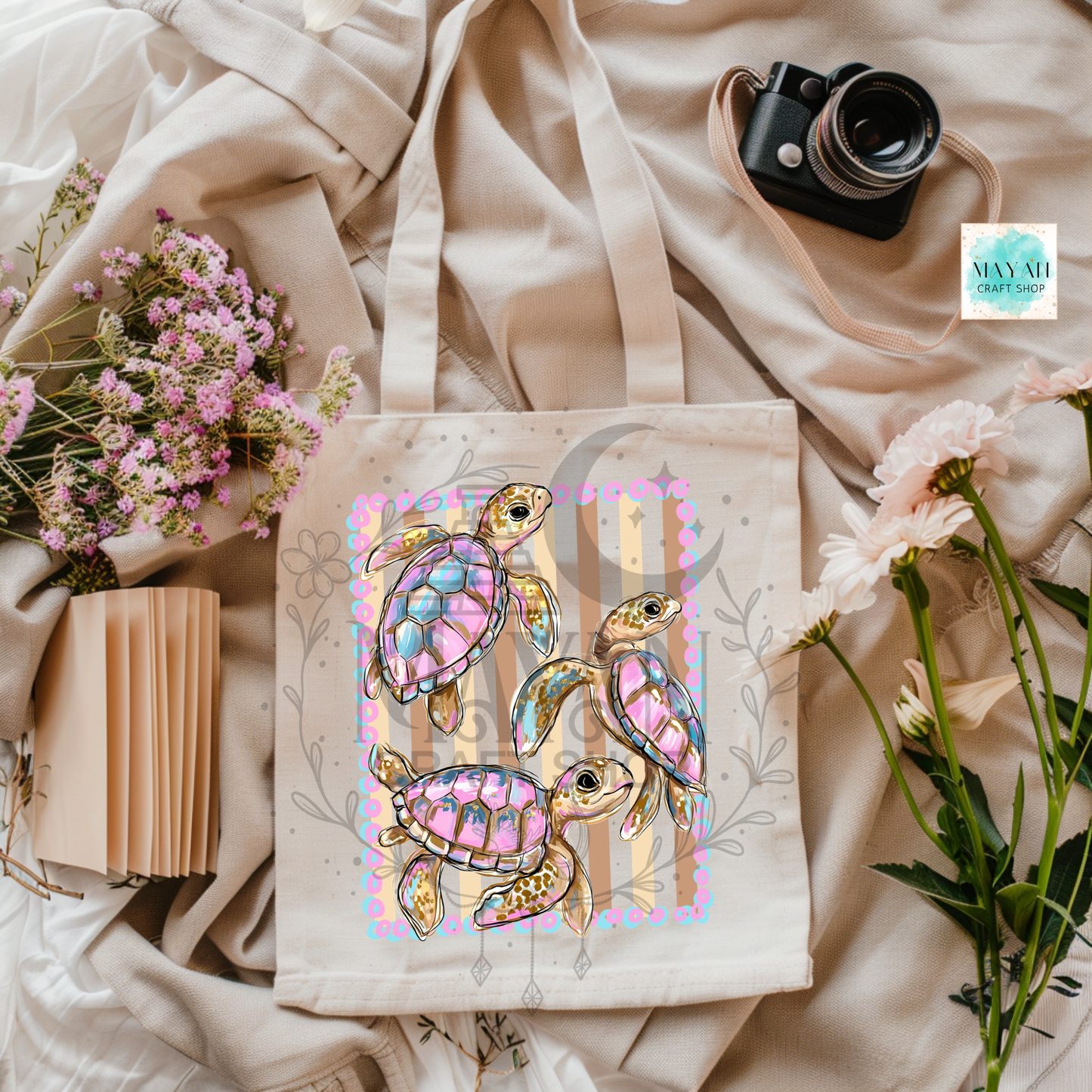 Tote bag with colorful turtle design on a soft fabric background with flowers and a camera.