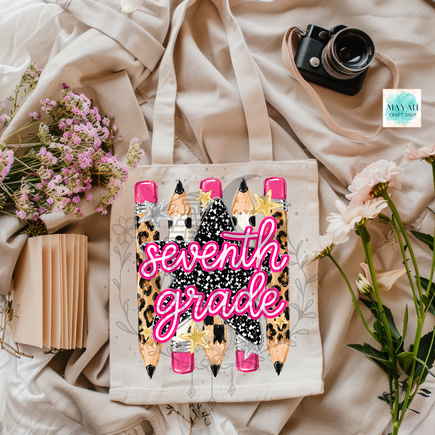 A canvas tote bag with a 'seventh grade' school-themed graphic featuring cartoon pencils and leopard print, styled in a flat lay with flowers, a camera, and a notebook on beige fabric.