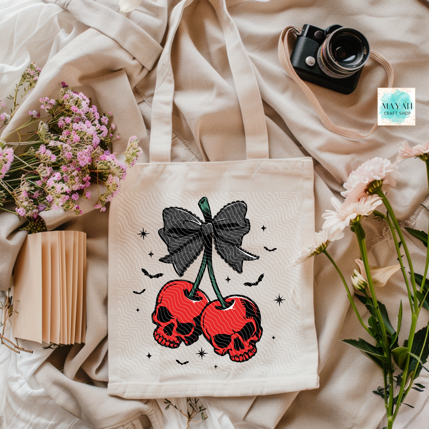 Tote bag with skull design on a soft fabric background with flowers and a camera.