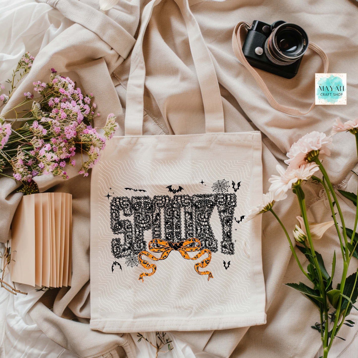 Tote bag with Halloween-themed design on a soft fabric background with flowers and a camera.