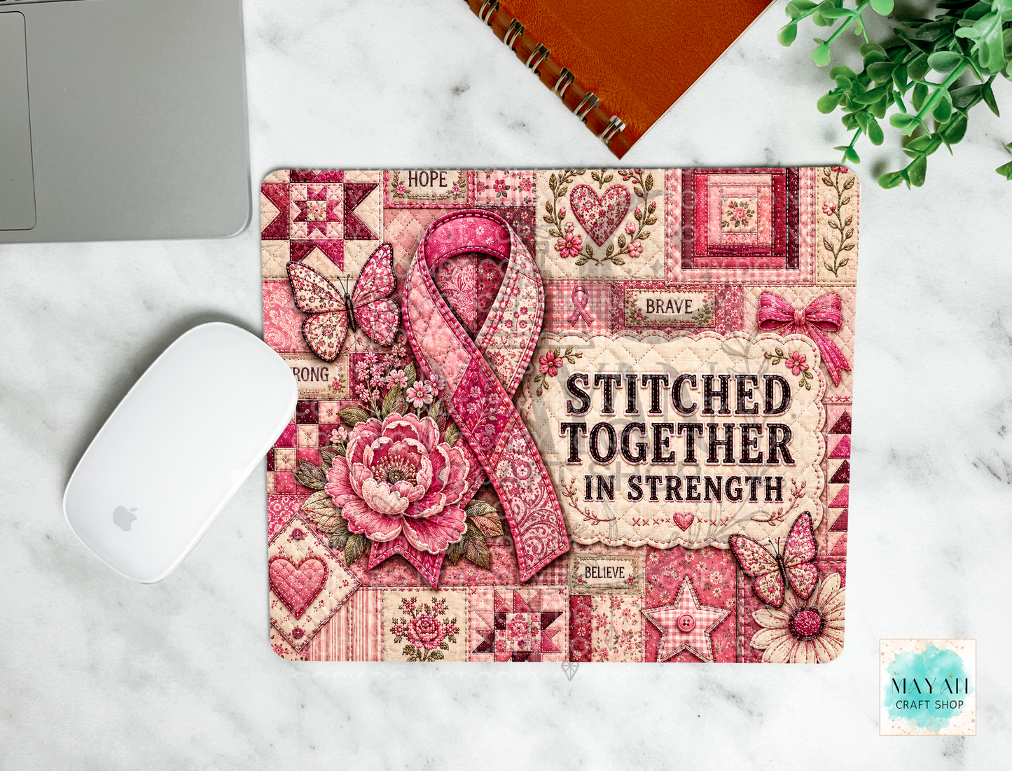 A rectangular mouse pad with a pink and beige quilted patchwork design, featuring a large pink ribbon, butterflies, flowers, and the text 'STITCHED TOGETHER IN STRENGTH'. The mat is displayed on a white marble surface next to a white computer mouse, a silver laptop edge, an orange spiral notebook, and a small green plant.